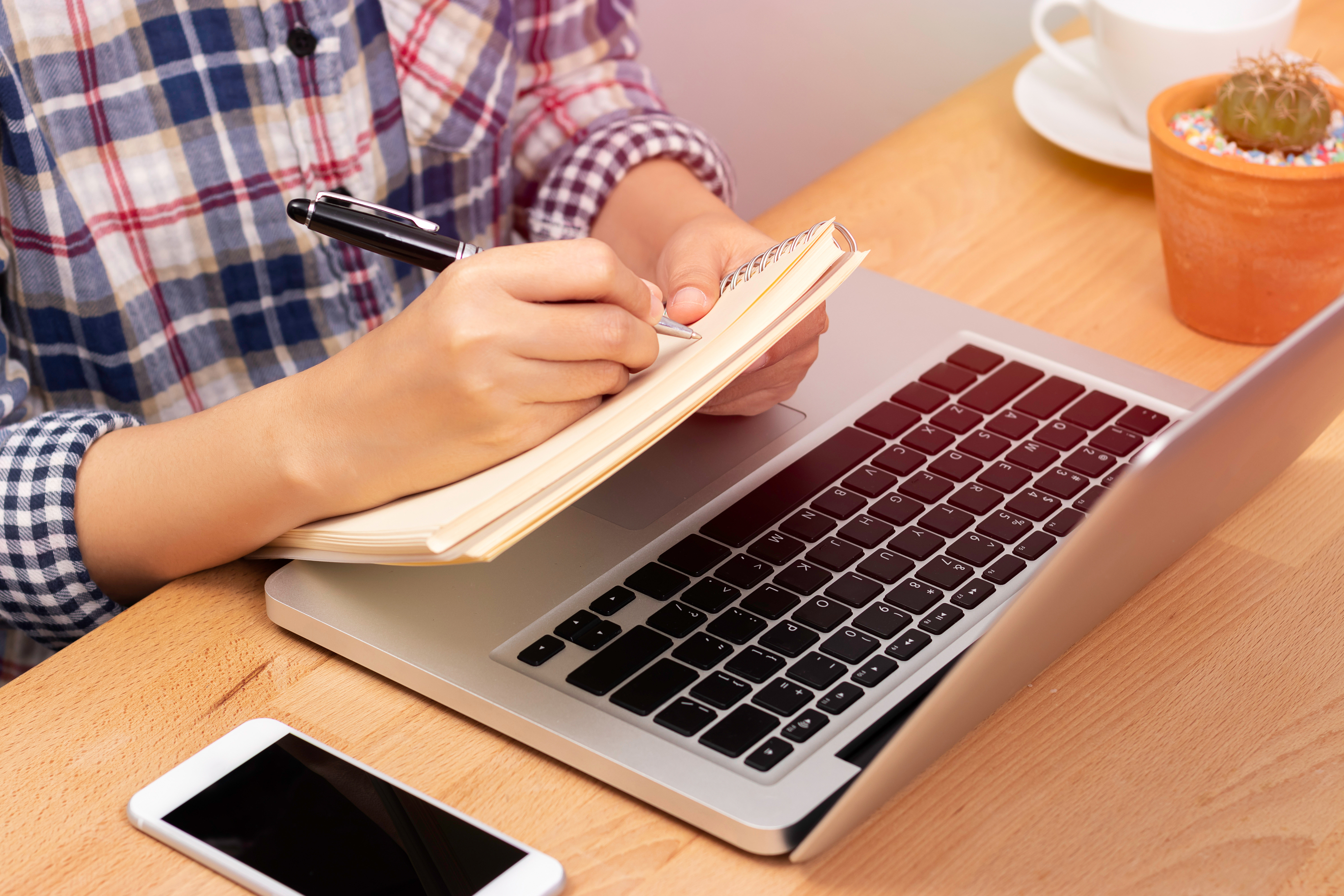 A person writing in a notebook with a laptop, phone, plant, and cup on their desk.