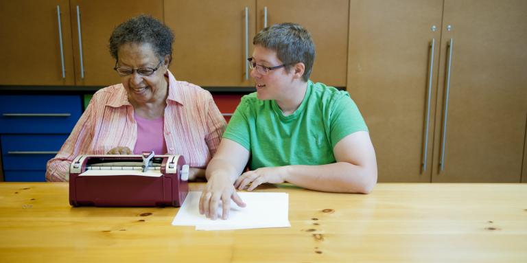 Older and younger woman sit at table in front of brailler 