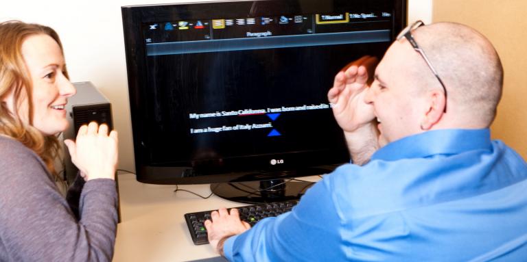 Man sits in front of office computer using magnification software, talking to woman 