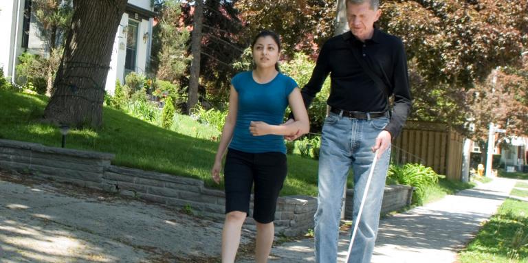 Older man and younger woman walk down sidewalk, man using white cane