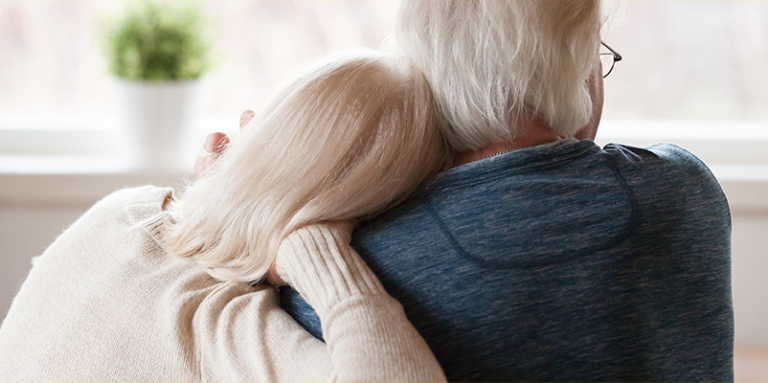 A man and woman sit on a couch facing away from the camera, comforting one another